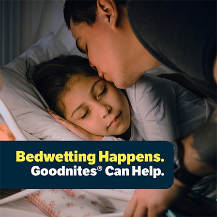 Parent kissing a sleeping child on the forehead at bedtime with title card saying: “Bedwetting Happens. Goodnites® can help.